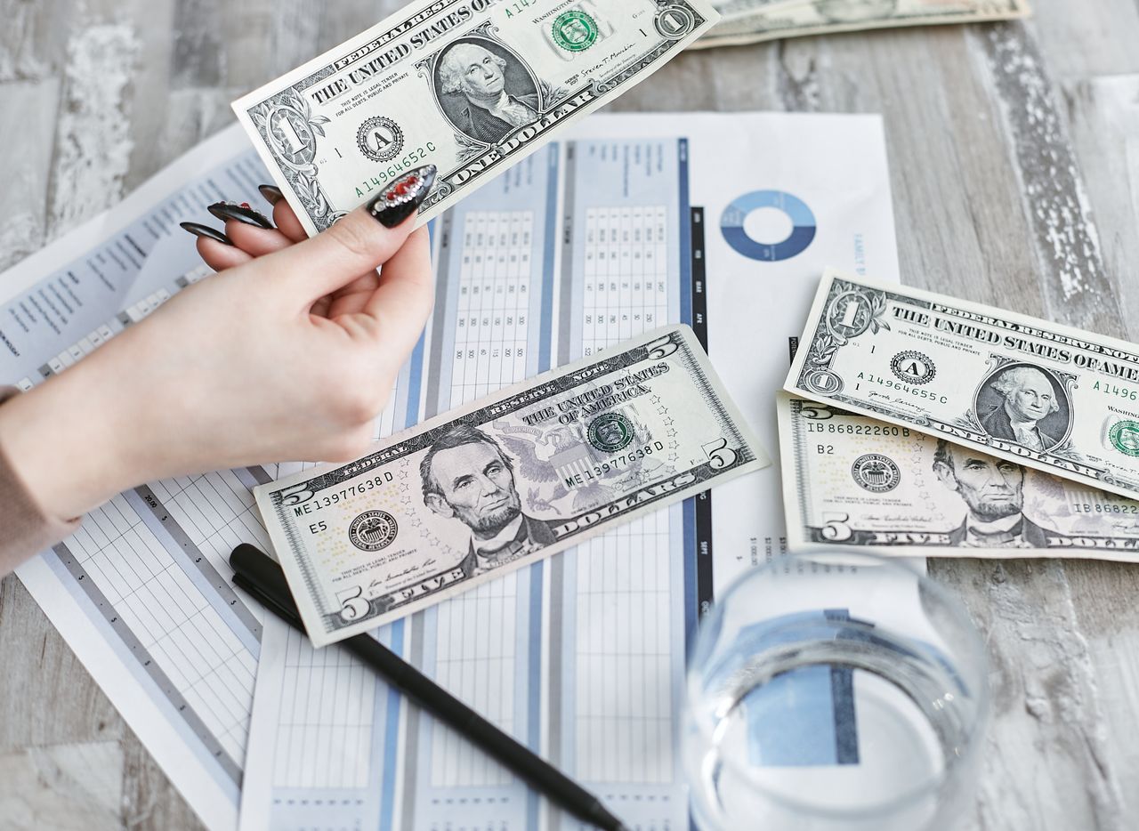 Close-up of a hand holding a dollar bill over financial documents and charts on a wooden table.